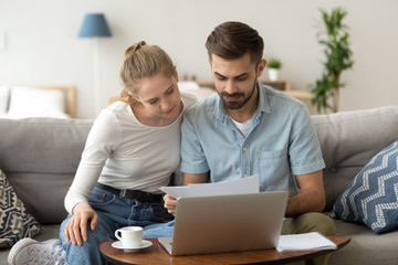 Young serious couple reading financial documents checking bills with laptop