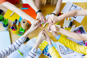 Fototapeta na wymiar Top view of hands of business people holding hands of each other over desk in creative office. Architects and interior designers at table with color samples, room layouts, supplies. Teamwork concept.