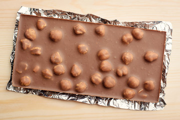 Bar of chocolate with many hazelnuts lays on foil on light wooden background. Top view.