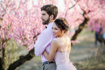 Newlyweds in love stand on the nature, against the background of wooden stakes, in sunny weather. Stylish groom embraces a beautiful bride in a lace dress in a green garden.