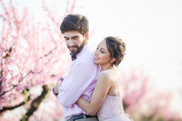 Newlyweds in love stand on the nature, against the background of wooden stakes, in sunny weather. Stylish groom embraces a beautiful bride in a lace dress in a green garden.