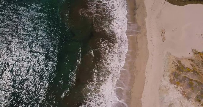 Aerial top view zoom out shot of beautiful foaming ocean waves reaching sunny exotic sandy beach in Big Sur California.