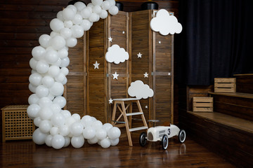 Birthday white decorations with balloons, stars, clouds and toy retro car for little baby on a dark brown background.