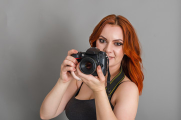 Smiling woman with red hair holds a modern camera in her hands. Gray background, creativity, photographer, beautiful