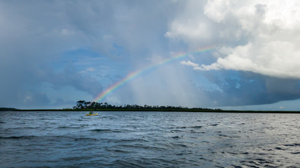 Boy paddling kayak across water looking at rainbow and clouds over distant shore
