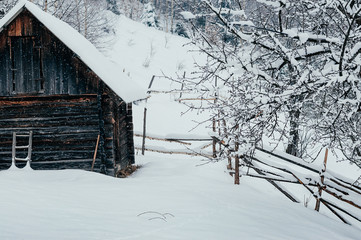 Winter countryside mountain landscape with wooden store house covered with snow, forests in the misty backdrop. Picturesque and peaceful wintry scene European resort location