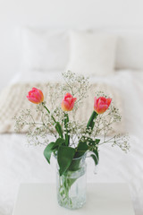 Glass vase with pink tulips and gypsophila flowers in light cozy bedroom interior. White wall, bed with white linen, beige thick yarn knitted woolen merino chunky blanket or plaid, pillows.