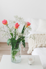 Fototapeta premium Glass vase with pink tulips and gypsophila flowers in light cozy bedroom interior. White wall, bed with white linen, beige thick yarn knitted woolen merino chunky blanket or plaid, pillows, candle.