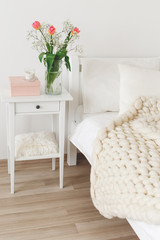 Cozy bedroom interior: white wall, bed with white linen, light beige thick yarn knitted woolen merino chunky blanket or plaid, pillows, bedside table, vase with tulips flowers.