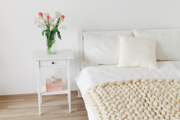 Fototapeta na wymiar Cozy bedroom interior: white wall, bed with white linen, light beige thick yarn knitted woolen merino chunky blanket or plaid, pillows, bedside table, vase with tulips flowers.