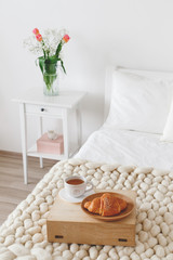 Cozy bedroom interior: white wall, bed with white linen, light beige thick yarn knitted woolen merino chunky blanket, bedside table, vase with tulips flowers, tray with a croissant and a cup of tea.