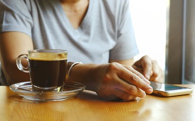 The hand of the woman who was scrolling for information on the screen on her mobile phone  and hot coffee in clear glass that she had prepared for drinking.