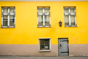 Fototapeta na wymiar Yellow dirty wall of a dilapidated house on a cobbled street of the old city of Riga. Wooden door with a cast iron bolt and windows randomly positioned on the wall. Facade.