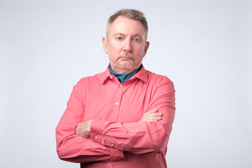 Serious senior man with folded arms and a deadpan expression posing on grey wall