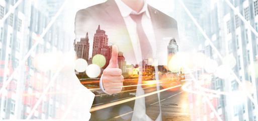 The double exposure image of the businessman wear black suit showing thumbs up overlay with cityscape. Concept of smart city, night life, business grow up.