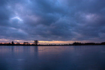 frozen lake, trees on the shore and clouds in the sky after sunset