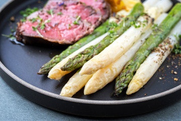 Traditional white and green asparagus with barbecue dry aged sliced beef fillet and fried potatoes served as closeup on a modern design plate