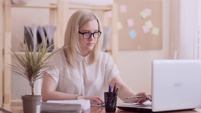 blonde girl caucasian appearance in glasses and white blouse left-handed in the office checks the documents on paper and computer on the table is a glass with pens and pencils.