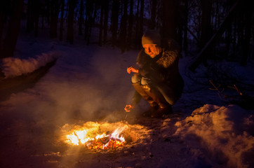Girl sit near bonfire roasting sausage on stick. Woman making food over fire in forest. Vacation and picnic concept. Roasted sausages food for hike picnic. Bonfire in the winter night forest