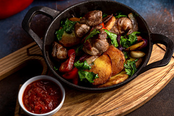 Frying pan with roast beef and potatoes, roasted vegetables and greens, on a light board on a black background. The background is decorated with vegetables.