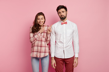 Horizontal shot of positive young woman with dark hair points at dissatisfied man who doesnt like formal festive clothes, ready for party, isolated over pink background. Look at my boyfriend