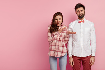 Photo of dissatisfied European woman demonstrates angrily at her male colleague, dressed in shirt, discontent with relationship, isolated over pink studio background with copy space aside for text