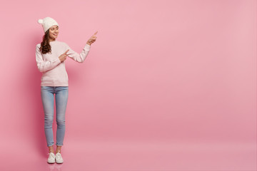 Studio shot of pleased European woman wears hat, jumper and jeans, shows direction aside, smiles gladfully stands against pink background with copy space for your advertisement or promotion.