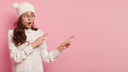 Astonished dark haired woman with curly hair points aside, has shocked expression, wears hat with ears and casual shirt, shows copy space for your promotional content or advertisement. Look there