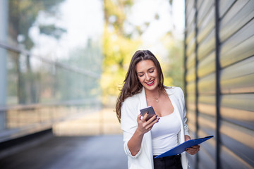 Fototapeta premium Young long hair woman in white dress using mobile phone with documents on hand near the office