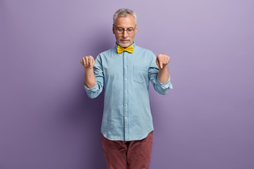 Serious bearded elderly man points down with both index fingers, shows product, promots copy space downwards against bright purple background, wears spectacles, stylish clothes. Look down please