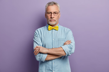 Fototapeta premium Horizontal shot of attractive woman with serious expression, looks directly at camera, keeps arms folded, wears yellow bowtie and denim shirt, isolated over purple background. Aging concept.