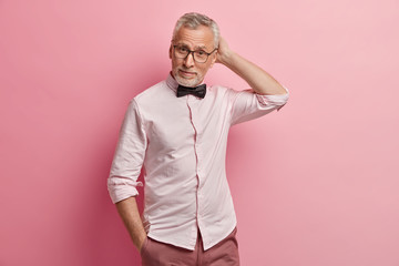 Waist up shot of attractive bearded grey haired man pensioner scratches head, holds hand in pocket,...