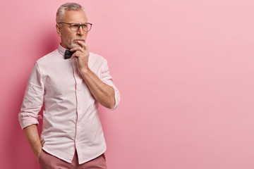 Pensive mature wrinkled man holds chin, lost in thoughts, looks away, keeps hand in pocket, dressed in formal clothes, models over rosy background with empty space for your promotional content