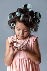 Funny cute little child girl in pink dress and hair curlers looks at the necklace