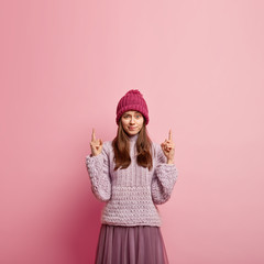 Photo of discontent woman purses lips, has angry expression, dressed in warm clothes, points upwards above, shows free space for your promotion, isolated over pink background. Look up, I dont like it