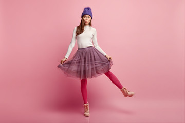 Studio shot of amazed pretty young woman demonstrates her new stylish skirt, wears bright pink...