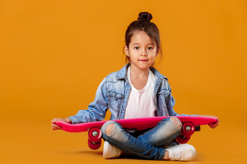 Naklejka premium Stylish little child girl with skateboard in jeans clothes on orange background