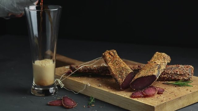 Pouring dark beer in glass and Jerky, basturma, dried meat beef, meat smoked jerky with spices on wooden cutting board, black concrete surface table background.