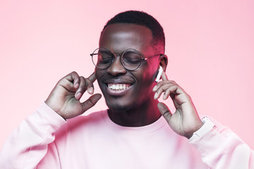 Studio shot of young african man listening to music with wireless earphones isolated on pink background