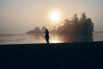 Silhouette of fisherman fishing with spinning rod on river bank at misty foggy sunrise. Isolated outdoor portrait of man standing by lake at sunset holding fishing rod and camp chair under his arm