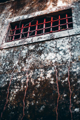 A vertical view of an old grungy wall with a rectangular window hole on the top and vivid red bars protecting it; the facade of an old house in Sintra, Portugal with red window, shallow depth of field