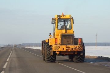 Big yellow bulldozer tractor rides along the asphalt road on the background of snowy fields and the blue of a clear sky in winter - rear view