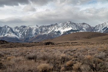 brown winter desert valley landscape with brown rolling hills, snowy mountain peaks of Sierra Nevada and gray cloudy sky in California, USA