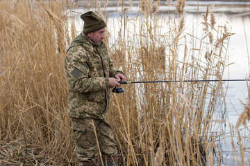 Fishing in the reeds for spinning on quiet water. A man in camouflage clothes in cool weather is fishing on the river bank.	