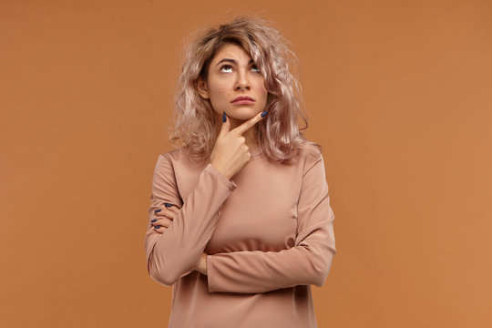 Pensive European female in her twenties making plans for weekend, rubbing chin and looking up, having thoughtful indecisive facial expression. Human emotions, life perception and body language