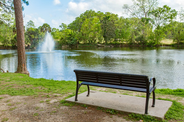 Fototapeta na wymiar Empty bench next to a lake with a fountain in Audubon Park, New Orleans, Louisiana, USA. Color horizontal photo, trees surround lake, fountain on left