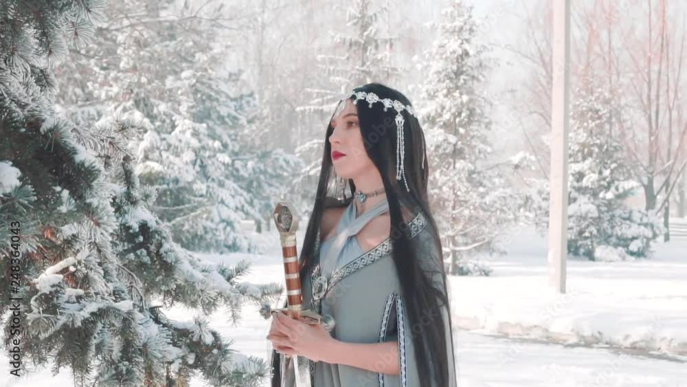 Wall mural woman fantasy warrior holding royal elven sword in hand. long black hair, red lips make-up posing for camera, elf princess with blade sharp weapon. Creative dress gray cloak in winter forest pine tree