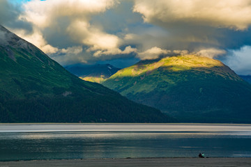 Golden light bathes the tips of mountains in Alaska overlooking an alpine lake. Striking cloud...