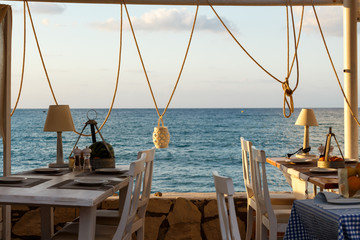 Seaside restaurant with white chairs and tables. Table set for dinner. Typical Greek seaside restaurant. Outdoor restaurant, taverna in Greece. View over the sea just before the sunset.