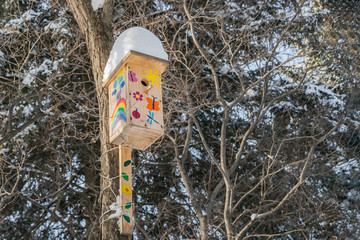 A birdhouse for birds from yellow boards with pictures and with white snow on the roof is on a brown tree in a park in winter